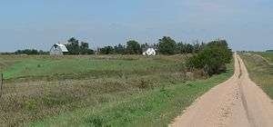 House and barn set in a line of trees in slightly rolling open country; gravel road runs by them