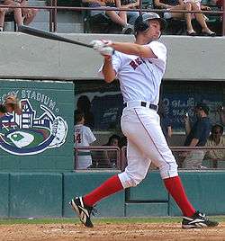 A man in a white baseball uniform and red socks swings left-handed at a baseball.