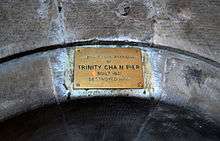 A colour photo of a brass plaque attached to greyish-pink stone masonry inside a building. The text on the plaque says: "Original Stone Anchorage/ of /Trinity Chain Pier/ Built 1821/Destroyed 1898