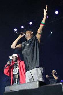 Three African-American men holding microphones stand on a stage; one man has his left arm raised up.