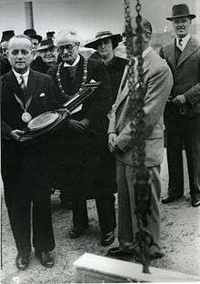 In a black and white photograph, a group of six men and two woman wearing mostly dark suits and mayoral decorations stand on a building site. A man on the far left bears a device resembling a large barometer.