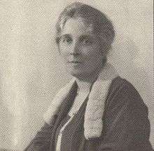 A seated woman, gray hair pinned up, in a black dress with fur collar before a plain white background