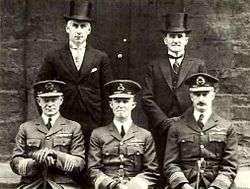 Portrait of five men, three seated wearing dark military uniforms with peaked caps, and two standing behind wearing formal suits with top hats