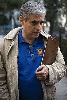 A middle-aged man in a blue polo shirt and khaki coat holds a paperboard.