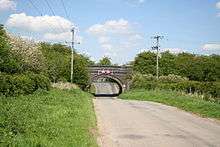 Blue brick single arch bridge carrying former railway over the road into the village.  The first houses are just visible through the arch.  The road surface has been lowered to pass under the bridge.  On either side steep embankments carry the former line, completely overgrown with trees and shrubs.