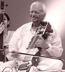 An old man sits cross-legged on a platform and plays a bowed instrument.