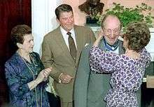 An elderly man is awarded a medal by a woman, she is attaching it around his neck whilst he grins excitedly.