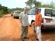 A photograph of two smiling men looking at the viewer and standing in the middle of a dirt road next to three automobiles under a blue sky