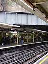 Eight people walking towards an escalator on a railway platform next to a green-tiled wall on the left and a railway track on the right