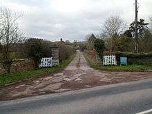 A road entrance to a building consisting of white-painted wooden gates, facebrick walling in a green landscape which prominently features various tree species.