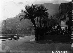 A large, open sports car drives past crowds of people and palm trees, with the city of Monaco in the background