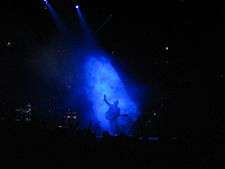 A man in a smoky blue spotlight singing at a microphone stand, holding a guitar in one hand and raising the other hand; off to his side and not in the light, another man standing and playing an electric keyboard