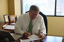 A grey-haired man with a white shirt and green tie sits at a desk in front of an open window, looking down to follow his pen as he writes on a sheet of paper