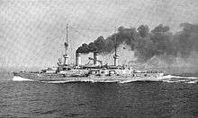 A large battleship plows through the water at high speed, thick black smoke pours from the smoke stacks