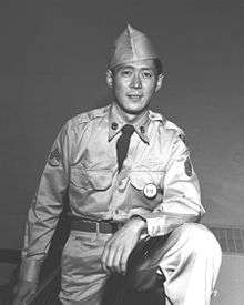 Young man in military uniform, standing with his arm resting on his raised knee.