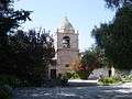 Photograph of the bell tower of Carmel Mission, with a peaked dome atop with gardens in the foreground.