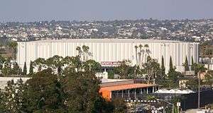 Outside view of stadium surrounded by trees and businesses; houses can be seen on large hills in the distance