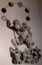 Raijin. Three-quarter view of a statue. His left leg is bend as if climbing stairs and he is leaning forward. His hair is standing up and his right hand is raised above his head. A thin halo around his head is decorated with thick circular disks. Black and white photograph.