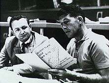 Two men seated at a desk, both reading from a large book the younger man is holding