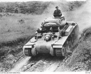 A tank cruises down an embankment in an open field while its commander stands out of the turret