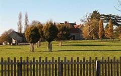 Stone buildings seen behind trees and grass area with wooden fence in the foreground.