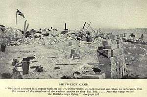 Boxes and equipment strewn around in disorderly piles on a broken ice surface. To the left a flag flies from a pole.