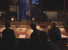 The inside of a music studio. Four men sit in front of a soundboard, while a woman wearing a red shirt stands inside the recording booth.