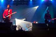 Two men playing a guitar in a concert, with another playing keyboards in the background.