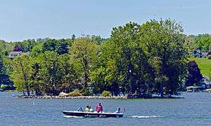 A boat with an outboard motor traveling across the foreground, in front of a small island in a larger body of water. Land rises up behind it.