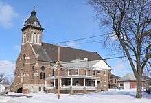 Two-story brick house in foreground; behind it, brick church with domed steeple and clocks in tower