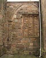 A dark painted wooden door, made of twelve vertical planks and dark iron hinges and nails, in an arched frame and wall of various light red and brown stones