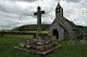 A plain greystone cross, in a mounting block, on top of four stone steps, with a small stone church in the background