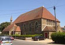 Three-quarter view of a long, low, brown brick and stone church with a high, red-tiled roof. An extension with two separate rooflines protrudes from the left side; it is topped by a small stone spire. There are three large windows on the same side.