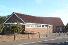 A long, low brick building in two non-aligned parts. Nearer the camera, in three-quarter view, is a hall with white soffits and window-frames; the windows are immediately below the roofline. Behind it, in profile and mostly hidden, is a longer building of brown and red brick and with a steep, high roof.