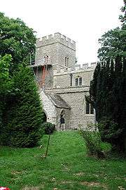 Seen between trees is part of the church; visible are a tower and nave with clerestory, both battlemented, and a porch