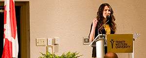 A photograph of a woman speaking into a microphone behind a golden sign that reads "TRINITY WESTERN UNIVERSITY" in black letters