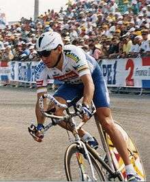 A man riding a bicycle wearing a helmet with a visor. Behind him there is a crowd of people on the road side