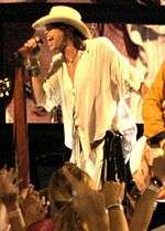 A man dressed completely in white, including a white cowboy hat, speaking or singing into a microphone. Another musician and several spectators are also partially visible.