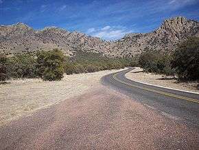 State Highway 166 winds toward Sawtooth Mountain in the distance. Thick growths of shrubs and small trees are along both sides of the road. Sawtooth Mountain has rugged outcrops of rock at its summit giving the mountain its name. Shrubs partially cover the mountains flanks.