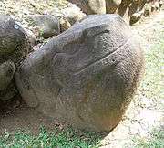 A smoothly finished boulder carved into the shape of a seated frog or toad, with the head raised to the right and sporting a prominent smile. The eyes, mouth, nostrils and legs are all carved in low relief. The sculpture is set against the stonework base of a structure, behind it at left.