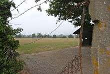 Through a wired fence, there is a grass playing field and a cricket pavilion to the right.