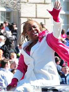 A woman is sitting and waving her hands. She is in her early sixties and wears a white suit and a pink scarf.