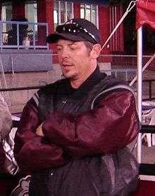 Half-length view of a person in his early 40s. He is standing upright with his arms folded across his chest. He is wearing a black and maroon coat and a baseball cap.