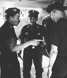 Three Asian men with black hair standing from left to right. The first is in profile, wearing a beret. He has three stars as a Lieutenant General and has a mustache. The next man is facing towards the camera smiling. He is clean shaven and has three stars. A third man on the right has his back to the camera and is wearing a cowboy hat. All are wearing military fatigues.