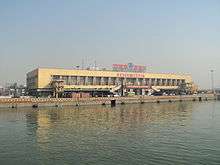 a long ochre low-rise building with broad windows. A pier extend parallel to the building