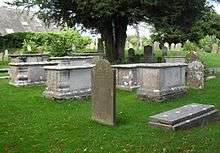 Three-quarter view of several large stone tombs and some gravestones in a grassed churchyard, with trees visible in the background.