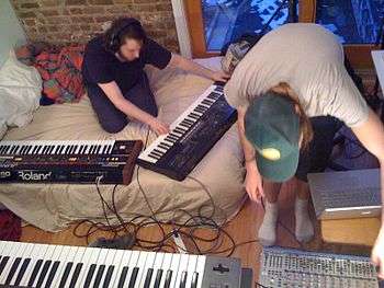 Musician with headphones playing a synthesizer, while another musician adjusts an equalizer