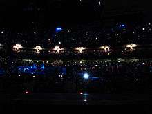 A darkened concert venue. People are in multiple levels of seating, and tiny lights are scattered throughout the audience. On the left, performers stand on a stage lit with blue lighting.