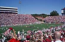 American football players line up for a kickoff on an American football field surrounded by crowded stands.