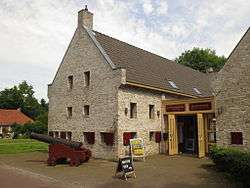 Light yellow brick building with dark red shutters and with a canon in front of it